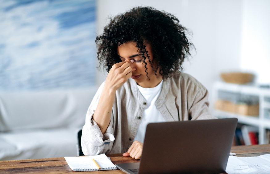Tired frustrated overworked African American young woman in casual wear, manager, freelancer or student sitting in living room at laptop, experiencing stress, headache, rubs and close her eyes with